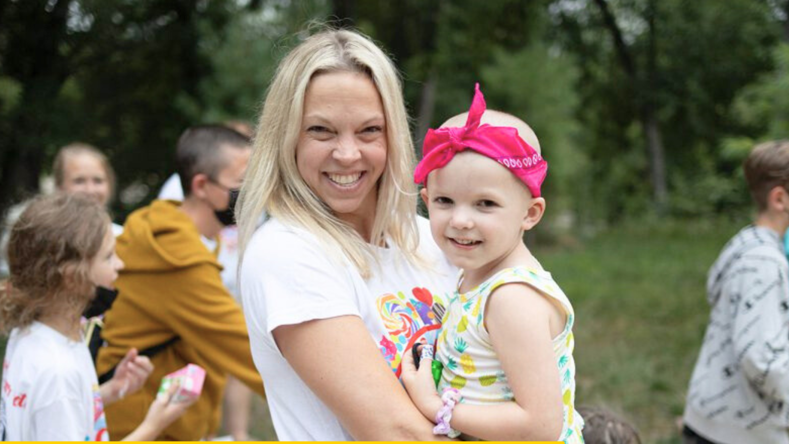 Mother holding a smiling child at a Care Camps Foundation event supporting children with cancer and their families during an outdoor camp experience.
