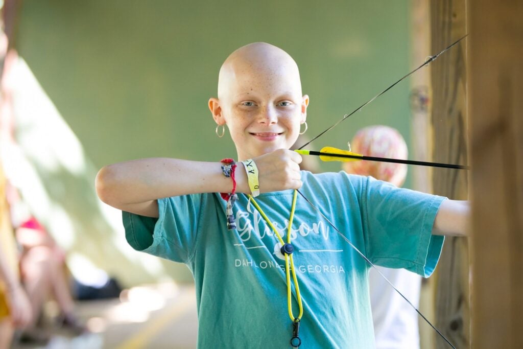 Child at Care Camps Foundation archery activity smiling while learning outdoor skills during a cancer support summer camp experience.