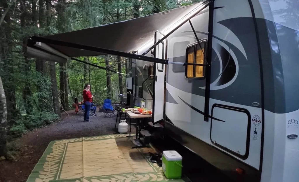 Travel trailer parked at a shaded forest campground campsite with the awning extended, outdoor chairs, and camping setup in Washington State.