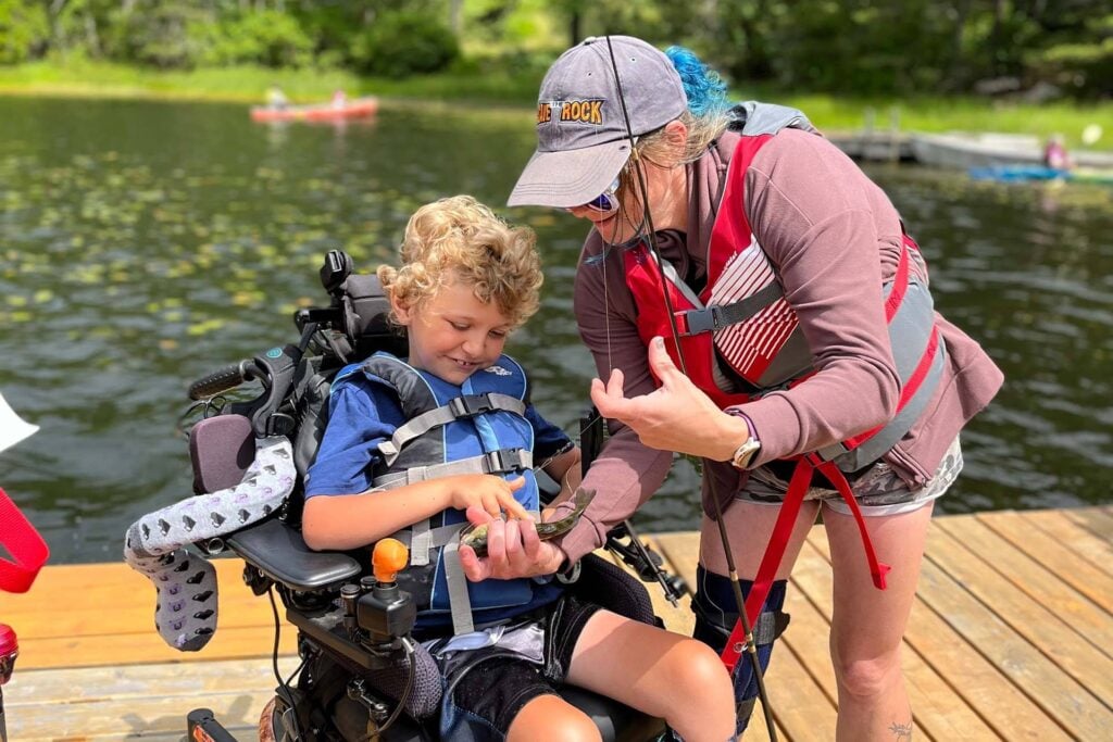 Child in a wheelchair fishing on a dock with assistance from a camp counselor during an inclusive outdoor camp experience supported by Care Camps Foundation.