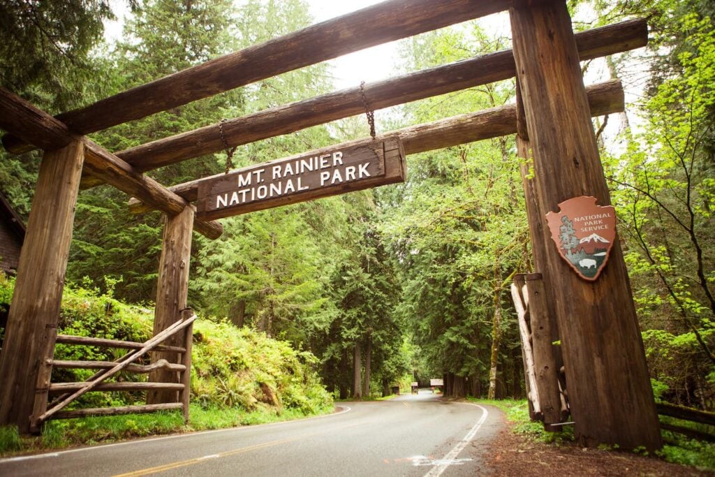 Mount Rainier National Park entrance sign surrounded by lush forest along a scenic Washington roadway.