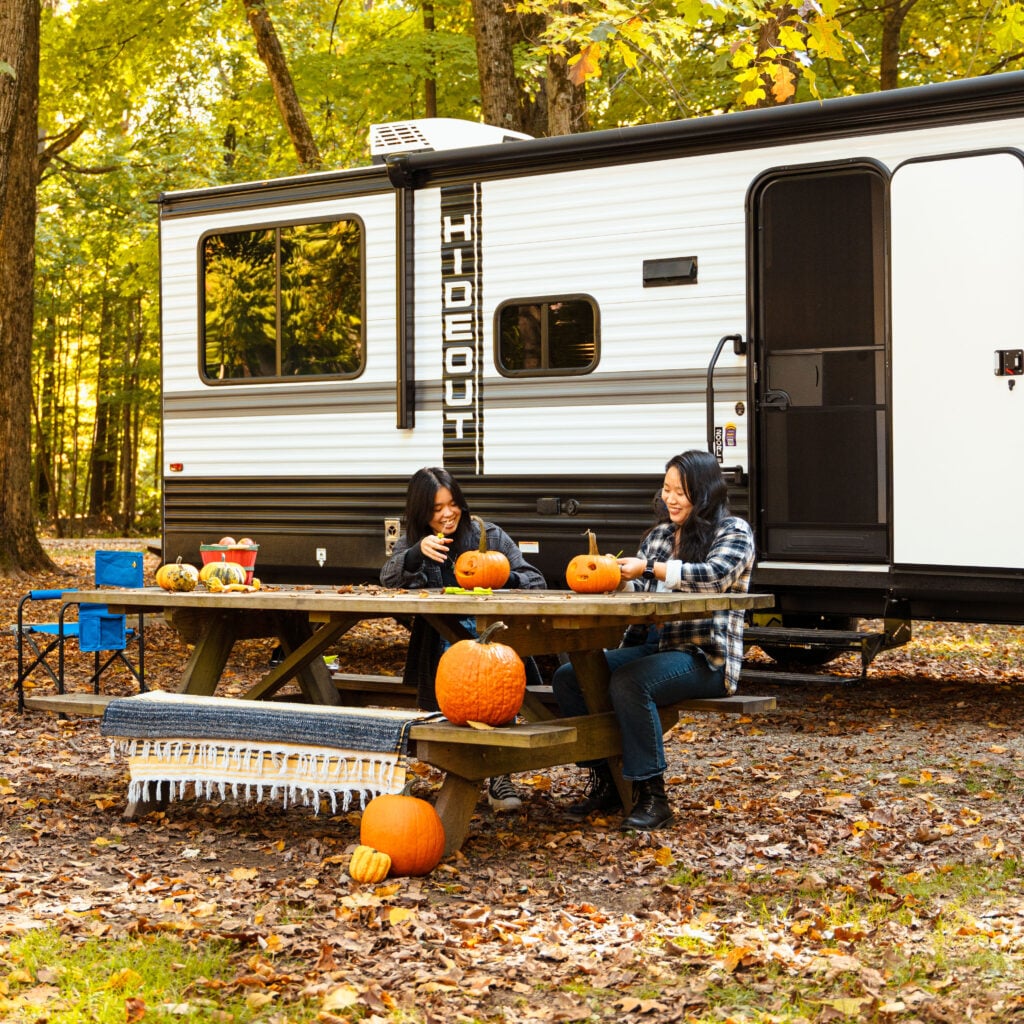 Friends carving pumpkins at a campsite next to a travel trailer in a wooded campground, highlighting fall RV camping in Washington state
