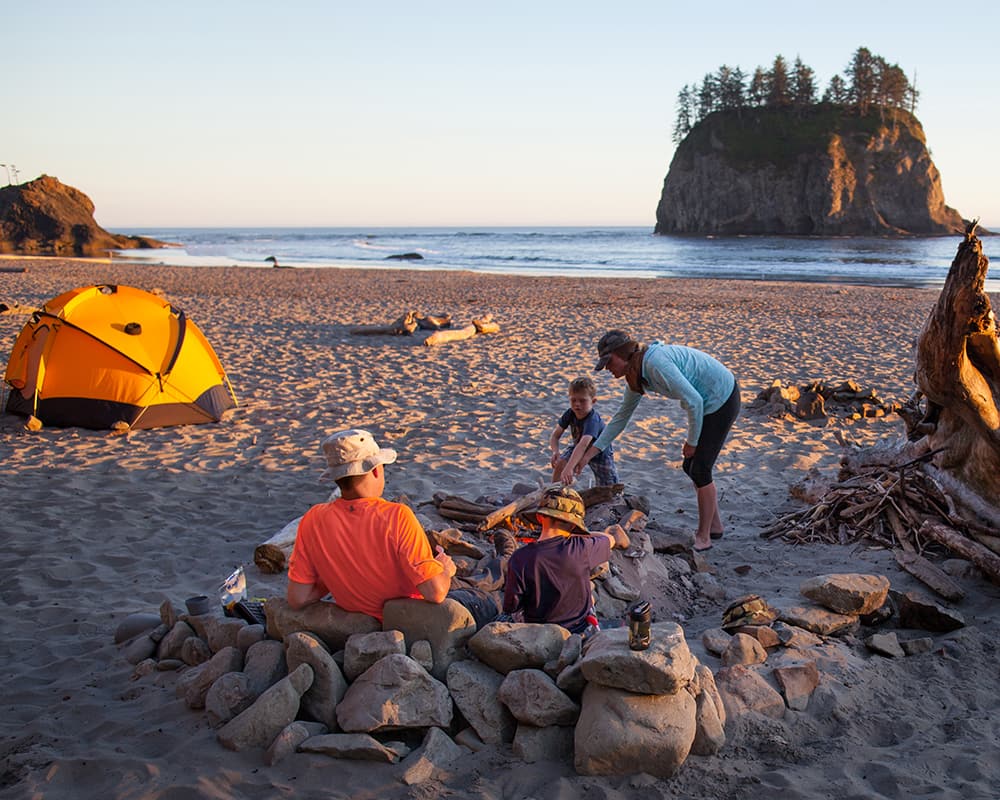 Family enjoying a beach campfire and camping adventure on the Olympic Peninsula coastline in Washington State during sunset.