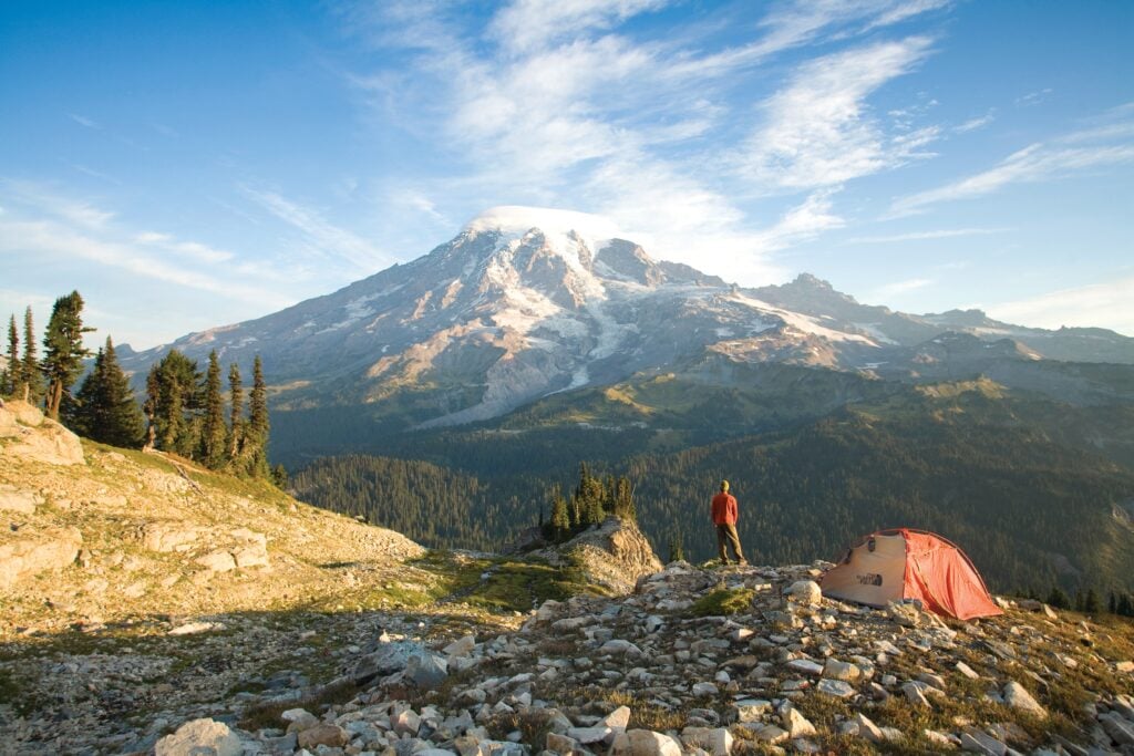 Hiker and tent overlooking Mount Rainier in Washington State during a backcountry camping adventure in the Pacific Northwest mountains.